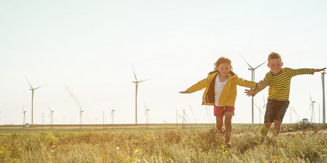 Eine Wiese auf dem zwei kleine Kinder rennen, im Hintergrund ist ein Windpark zu sehen